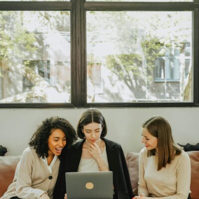 Three diverse businesswomen sitting together on a couch, collaborating on a laptop in a bright office setting.